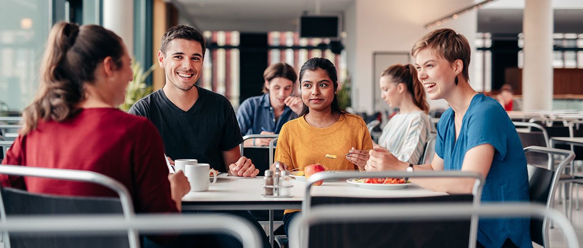 vier studierende sitzen am tisch einer mensa. sie essen und unterhalten sich angeregt.