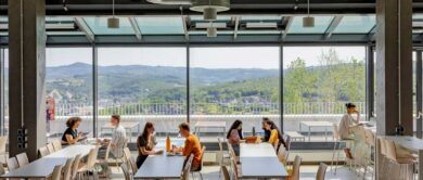 in einer mensa vor einer schönen talkulisse sitzen acht studierende an tischen und nehmen ihre mahlzeiten ein und unterhalten sich angeregt miteinander oder arbeiten an ihren laptops