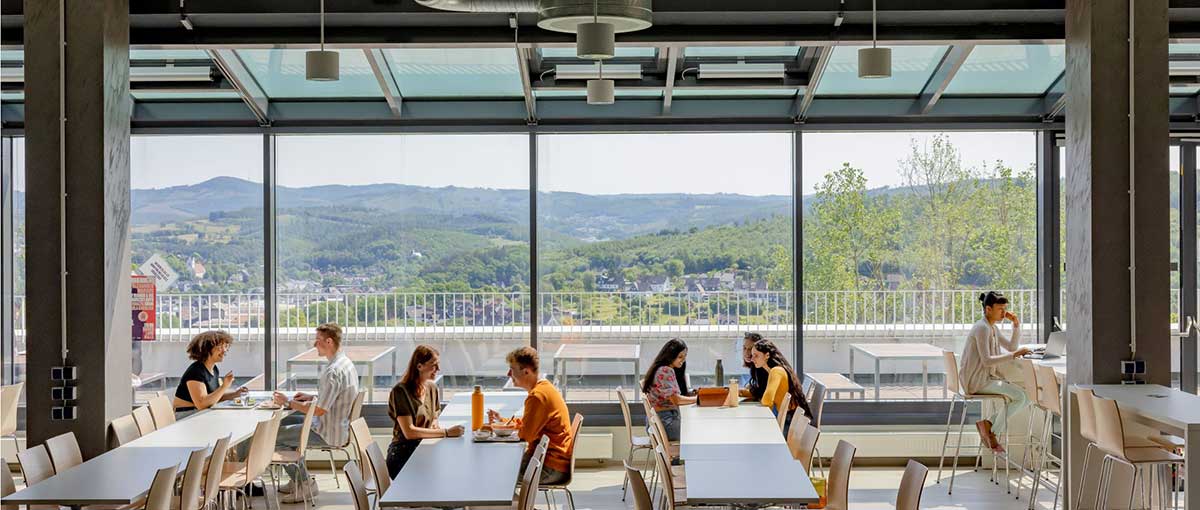 in einer mensa vor einer schönen talkulisse sitzen acht studierende an tischen und nehmen ihre mahlzeiten ein und unterhalten sich angeregt miteinander oder arbeiten an ihren laptops