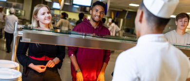 drei studierende (eine studentin und zwei studenten) stehen vor der ausgabetheke einer mensa und geben ihre bestellung auf