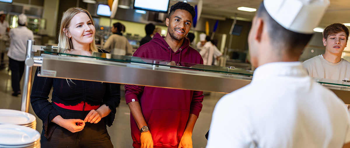 drei studierende (eine studentin und zwei studenten) stehen vor der ausgabetheke einer mensa und geben ihre bestellung auf